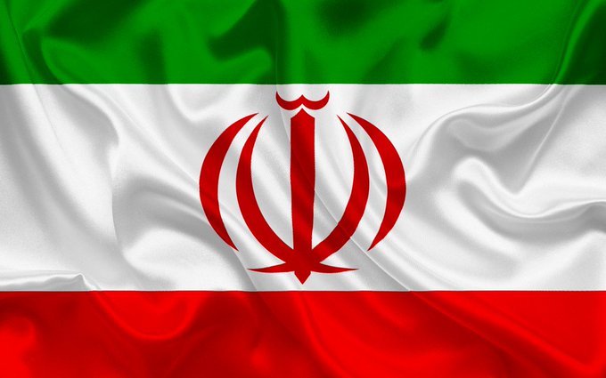 The image features a close-up view of a flag with red, white, and green colors. The flag appears to be waving or fluttering due to its texture. The background of the image is blurred, making it difficult to determine what lies beyond the flag. The caption provided in the image reads "BREAKING: US sanctions Iran'S largest Bitcoin and crypto exchange Nobitex over alleged IRGC and Central Bank links." This text seems unrelated to the flag's colors or waving motion but might be a part of an article discussing the impact of US sanctions on Iran's financial industry.