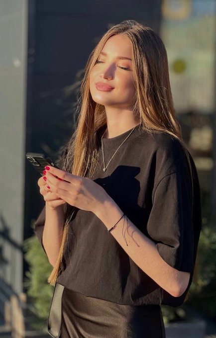 A young woman with long blonde hair standing outdoors in golden sunlight, smiling with her eyes closed while looking at her phone, wearing a black t-shirt and a delicate necklace.