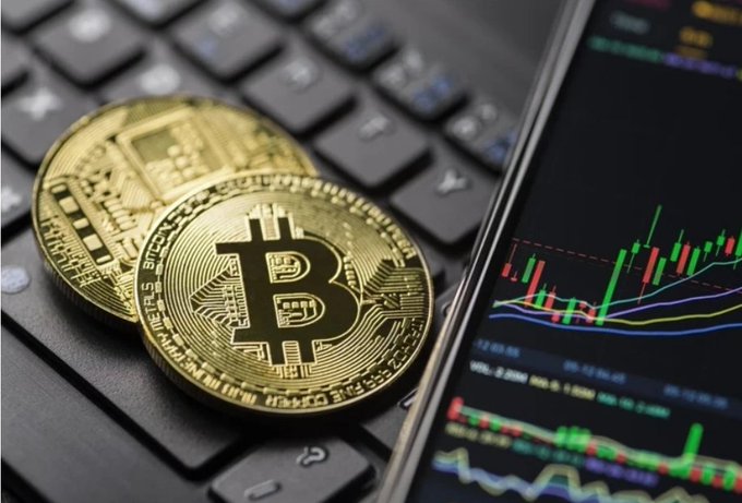 Two gold Bitcoin coins resting on a computer keyboard next to a smartphone displaying a financial trading chart with red and green candlesticks.