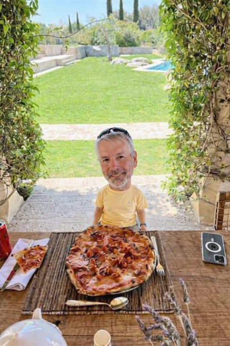 A man with sunglasses on his head sits outdoors at a table with a pizza topped with meat, with a garden and pool visible in the background.