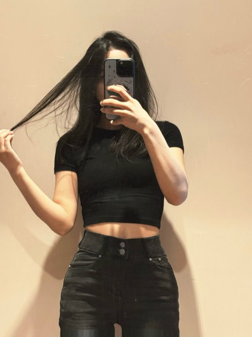 A woman takes a mirror selfie while wearing a black crop top and black high-waisted jeans, holding a strand of her long dark hair.