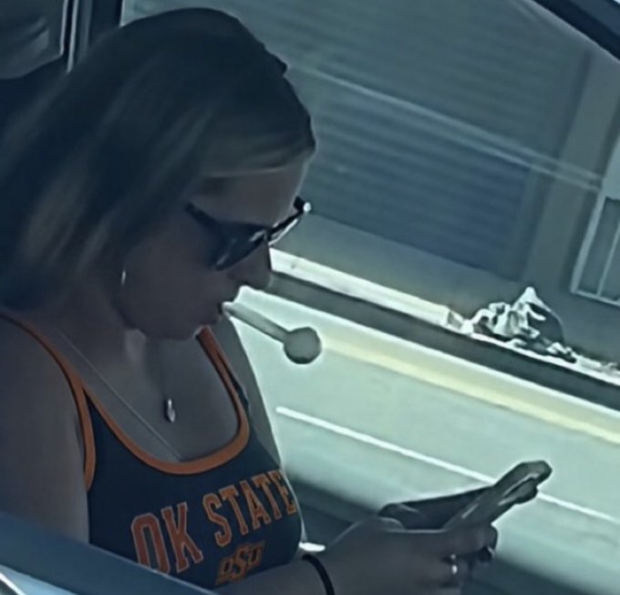 A screenshot from a dashcam showing a woman wearing sunglasses and an Oklahoma State University tank top looking at her phone while driving.