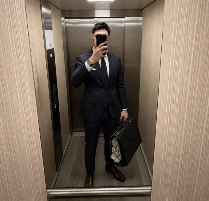 A man in a dark pinstripe suit takes a mirror selfie with his phone while holding a black briefcase full of US dollar bills inside an elevator.