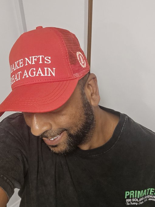 A man wearing a red cap that reads 'MAKE NFTS GREAT AGAIN' and a dark shirt with 'PRIMATE 000 SOLANA AVENUE' text, looking downward.