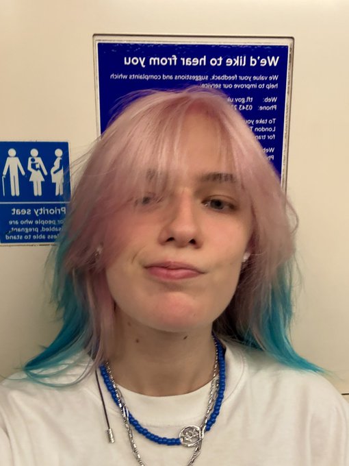 A selfie of a smiling person with shoulder-length hair dyed pink and teal, wearing layered necklaces, posed in front of inverted 'Family Bathroom' and 'Toy Centre' signs on a wall.