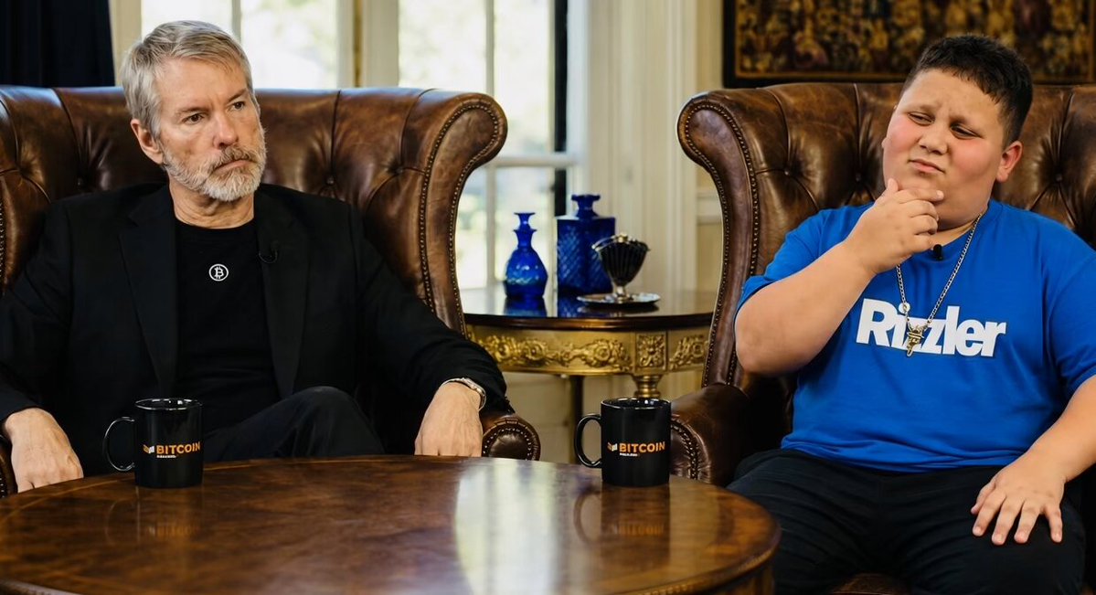 The image shows two men sitting at a table, with one man wearing a black shirt and the other wearing a blue shirt. They are both holding coffee cups, suggesting they might be having a conversation or discussing something related to Bitcoin. The background features a brown leather chair on which the men are seated, along with a vase and a bottle placed nearby. There is also a book visible in the scene.