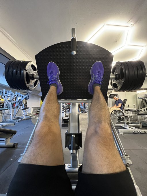 First-person POV photo of someone at a gym using a leg press machine, wearing purple sneakers, with another person visible working out in the background.