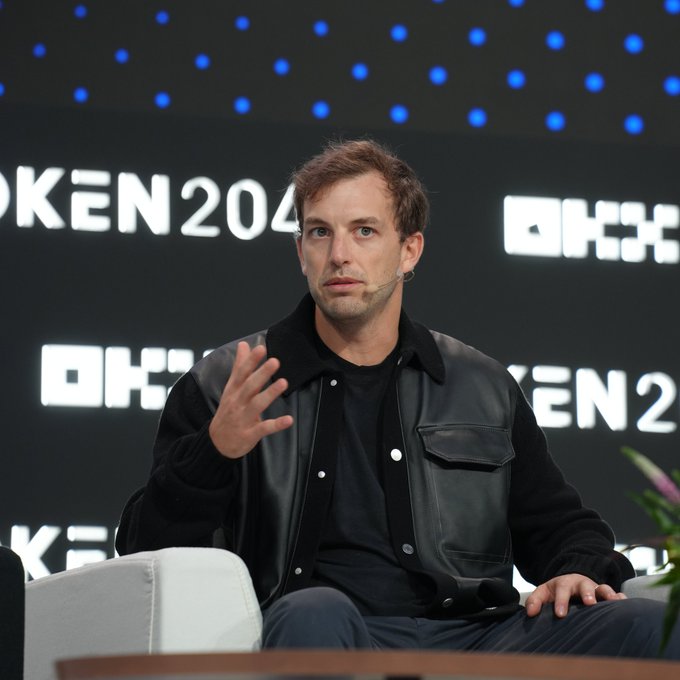 A man in a black leather jacket sits on stage at the TOKEN2049 conference, gesturing with his hand while speaking.