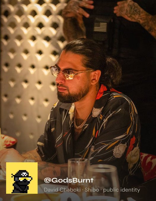 A man with a beard, glasses, and long hair tied back, wearing a black floral shirt, sitting at a table; overlay text reads '@GodsBurnt' and 'David Chaboki · Shibo · public identity' next to a pixel-art character logo.