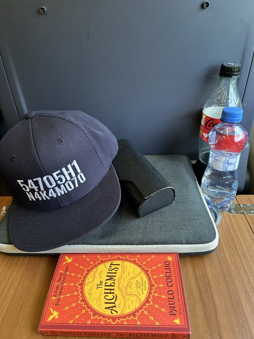 The image features a book titled "The Alchemist" by Paulo Coelho, which appears to be placed next to a bottle of water and a hat with the number 547506101 printed on it. The book's cover design includes an illustration of a map and a yellow bird. The post about Bitcoin is located in the bottom right corner of the image.
