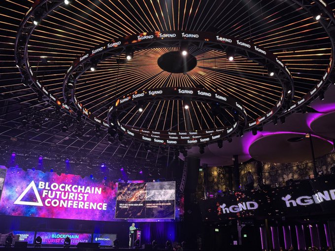 An ornate circular ceiling with radiating lights at the 'Blockchain Futurist Conference' venue, featuring 'GRND' branding and a large screen displaying the conference logo.