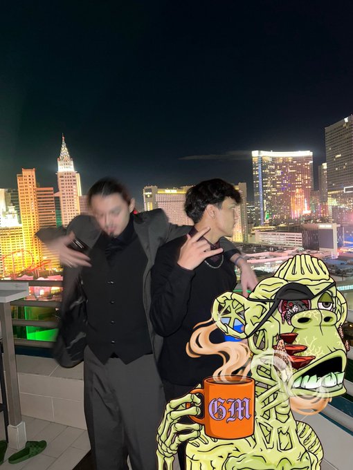 A nighttime photo of two young men in dark clothing posing on a rooftop with a brightly lit city skyline behind them, featuring a skeletal zombie character in the foreground holding an orange mug labeled "GM".