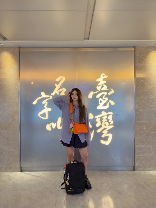A young woman posing in front of a wall with illuminated Chinese calligraphy, wearing a purple shirt and carrying an orange bag.