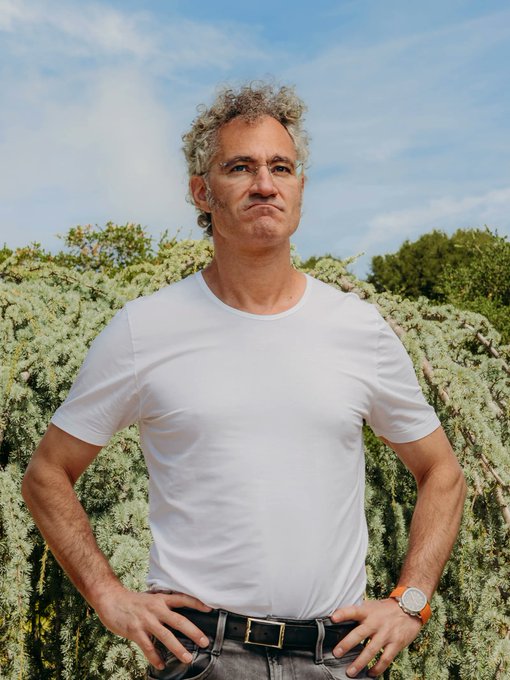 A man with curly graying hair and glasses, wearing a white t-shirt and a watch, standing outdoors with his hands on his hips against a backdrop of green bushes under a blue sky.