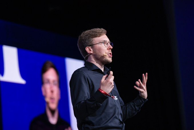 The image shows a man standing on stage giving a speech, wearing glasses and a black shirt. The background features a large screen displaying an image of a man's face with the text "I really think Ethereum's best time is yet to come." This suggests that the speaker might be discussing the potential impact or future developments in the world of cryptocurrency, specifically Ethereum.