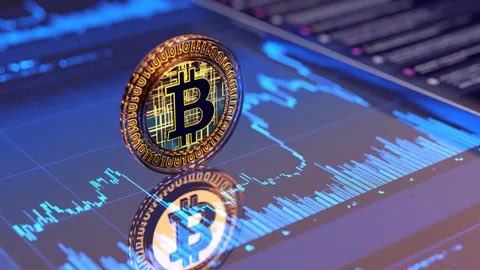 A close-up photo of a physical Bitcoin coin standing upright on a surface displaying blue-lit cryptocurrency price charts, with its reflection visible below.