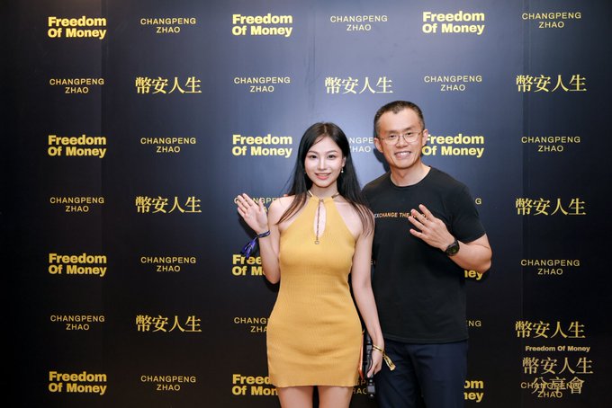 A young woman in a mustard yellow dress and a man in a black "Exchange the World" t-shirt pose together in front of a step-and-repeat backdrop featuring "CHANGPENG ZHAO," "幣安人生" (Binance Life), and "Freedom Of Money" branding. The image captures their meetup at the event, visually connecting to the post's celebration of attending the "幣安人生" gathering and meeting CZ (Binance's founder).