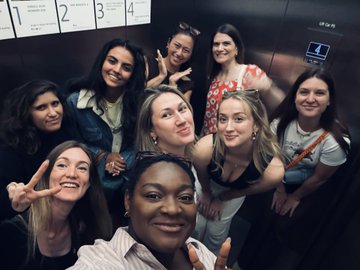 A cheerful selfie of eight young women posing closely together inside an elevator, with several making peace signs, capturing a fun and unified moment. Visible text on the wall shows floor numbers "2, 3, 4" alongside what appears to be an "air shower" notice, complementing the post's theme of bridging communities together at the Tate Modern in London.