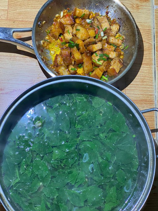 The image shows a table with two pots of food, one containing green vegetables and the other filled with meat. The vegetables are steaming hot, indicating that they have been cooked recently. The pot with the vegetables appears to be larger than the one with the meat. The scene suggests that someone is preparing a meal or a dish using both vegetables and meat as ingredients.