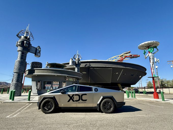 A silver Tesla Cybertruck with "XDC NETWORK" branding is parked in front of a retro-futuristic, UFO-shaped diner and space-themed structures under a clear blue sky.