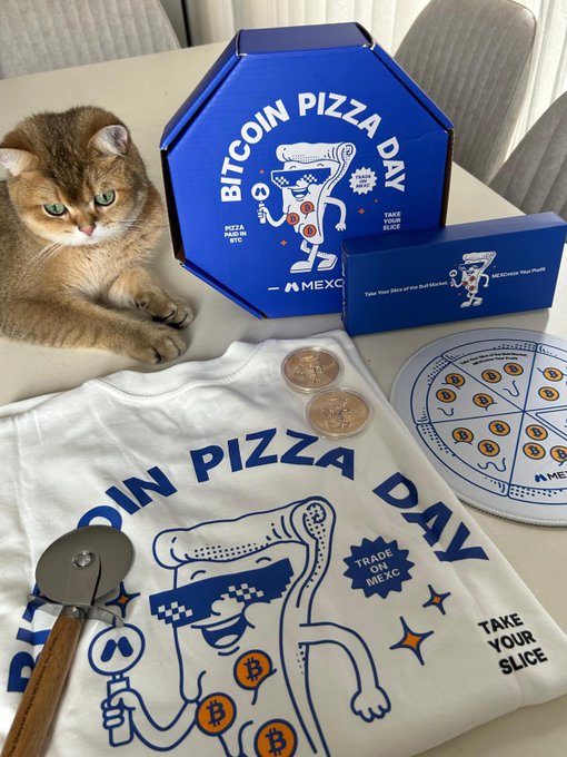 The image features a cat sitting next to a pizza box, with a slice of pizza placed nearby. The pizza box contains a pizza cutter and a pizza pan, along with other items related to a Bitcoin Day celebration. A mouse wearing sunglasses can be seen in the scene as well. The text on the post mentions that it was created by @MEXC and @SouBolado, which are likely the creators of the Bitcoin Day box.