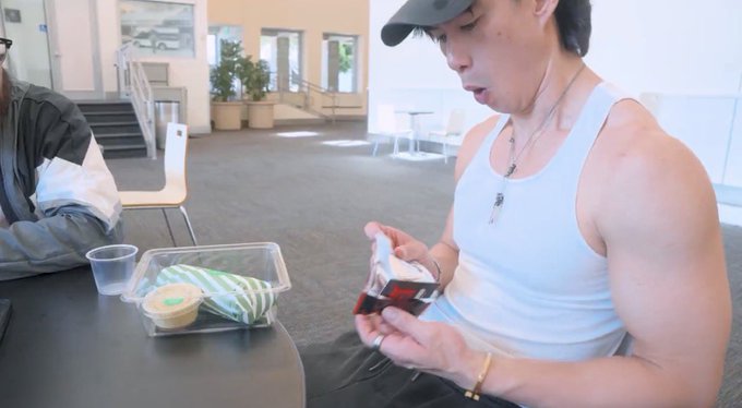 A man in a white tank top and black cap sits at a table examining a trading card from a clear plastic pack, with a takeout container and dipping sauce beside him. The scene captures the moment he's opening his Azuki TCG Regionals pack, moments before discovering the rare chase card inside.