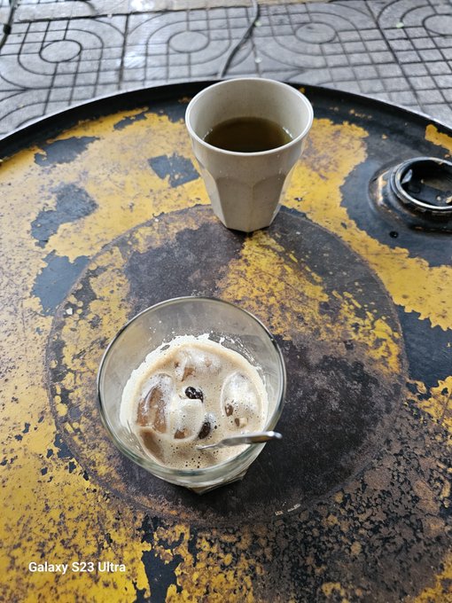A small cup of dark coffee and a glass bowl of iced coffee with milk and sugar sit on a weathered yellow and black metal tray on a tiled floor.