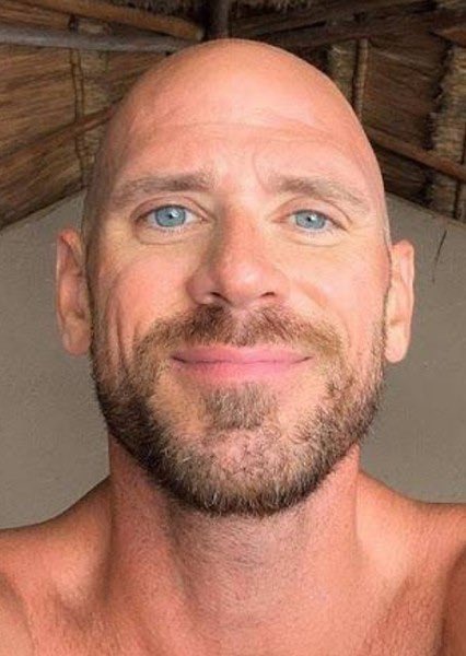 Close-up selfie of a bald man with blue eyes and a short beard, appearing shirtless against a wooden background.