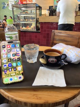 A first-person view of a cafe table holding an iPhone with various app icons, a glass of water, an empty espresso cup on a saucer, a water bottle, and a napkin.