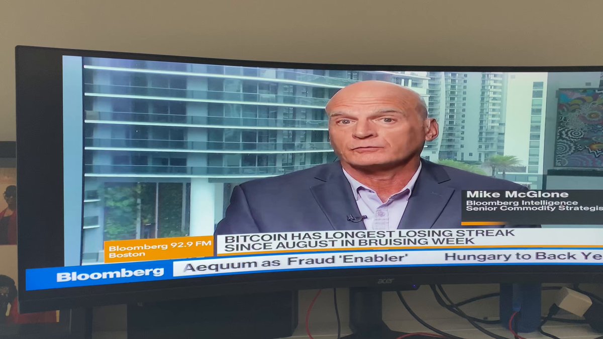 The image shows a television screen displaying a news program with a man wearing a suit talking about Bitcoin. The TV is placed on top of a desk, and there are two people visible in the scene - one person sitting at the desk and another standing near the TV. The focus of the image is on the man speaking into the camera, who appears to be discussing Bitcoin or its implications.