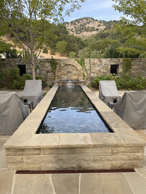 Stone reflecting pool with a fountain in an outdoor courtyard setting, surrounded by covered furniture and stone walls, with a hillside landscape in the background.