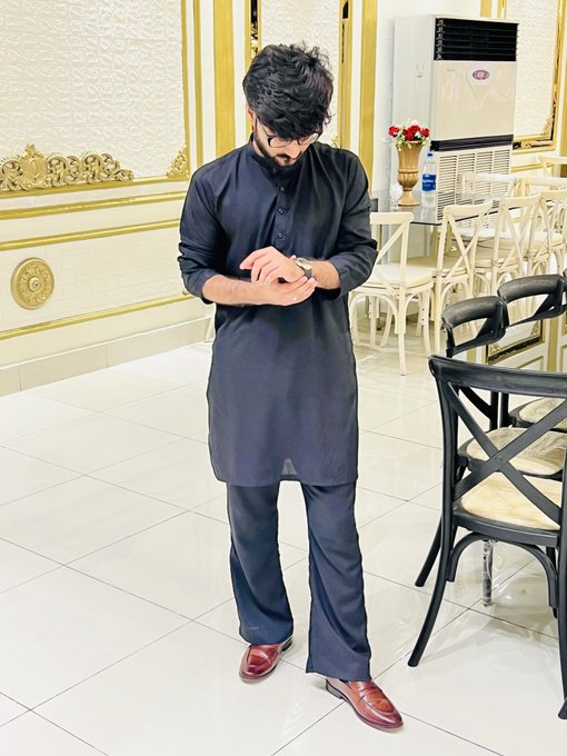 A young man with dark hair stands in an ornately decorated room with white and gold walls, looking down at his watch while wearing a long black traditional tunic (kurta) and brown shoes.