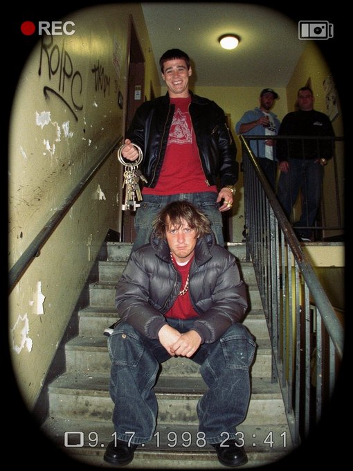 A digicam-style photo dated 9.17.1998 showing two young men on a graffiti-covered stairwell, one standing and one sitting, both wearing puffy jackets.
