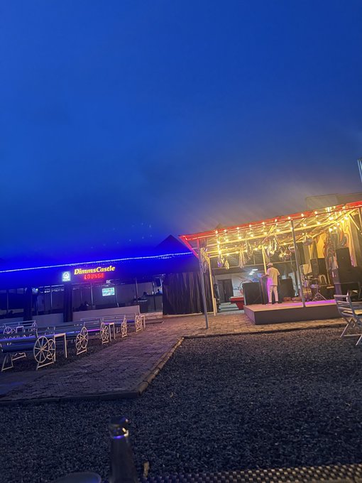 An evening outdoor scene featuring a stage with a performer under string lights and a 'Blimsa Castle' sign on an adjacent structure, set against a deep blue sky.