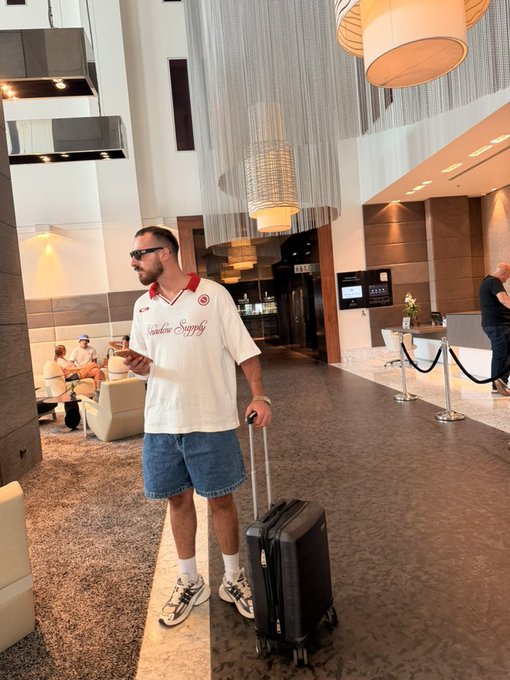 A man wearing sunglasses and a white "Shadow Supply" jersey stands in a modern hotel lobby holding a drink and the handle of a black rolling suitcase. The "Gm friends" caption reflects his relaxed, well-dressed traveler vibes as he seemingly checks in or out.