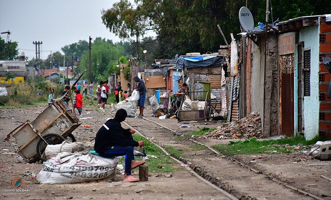 A photograph by Dado Soldani showing a person in a black hooded jacket sitting on a sack in a poor neighborhood with railroad tracks, makeshift houses, and other people walking in the background.