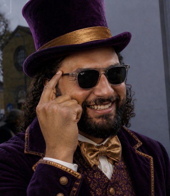 A man wearing a purple velvet top hat and matching coat with a gold bow tie adjusts his sunglasses while smiling, evoking the style of Willy Wonka.