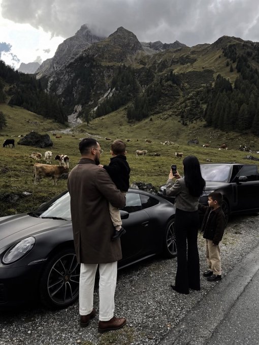 A family poses next to a black sports car and a black pickup truck on a mountain road, with grazing cows and majestic, cloud-covered peaks in the background.