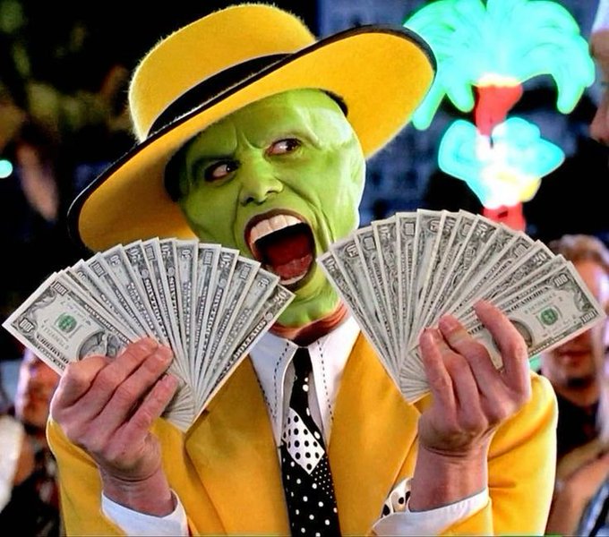 The Mask from the movie 'The Mask' is shown with a green face, yellow suit and hat, holding up two fans of cash bills with an excited expression.