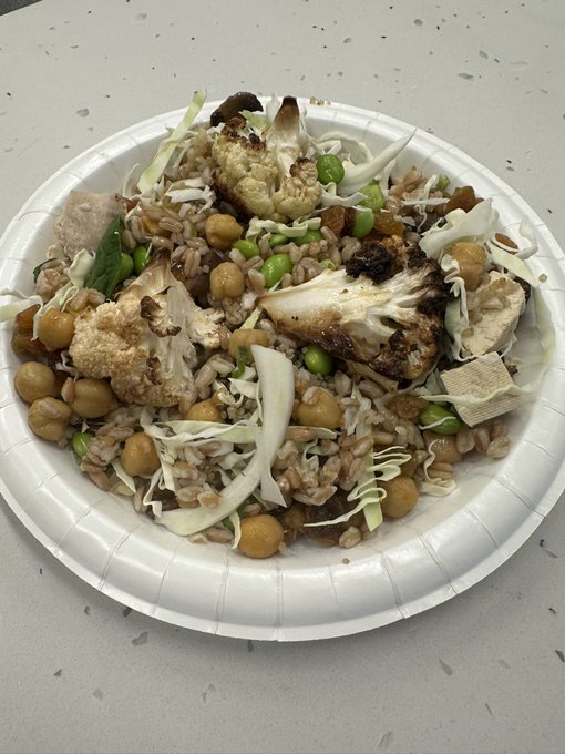 A white paper plate filled with a grain bowl containing farro, roasted cauliflower, chickpeas, edamame, tofu, and shredded cabbage.
