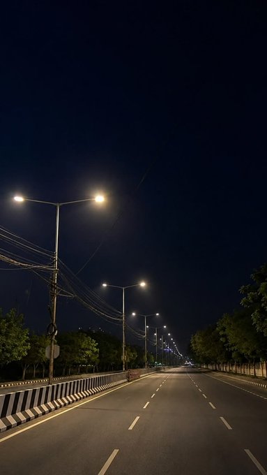 A nighttime photograph of an empty, well-lit multi-lane road lined with streetlamps, trees, and a black-and-white striped median barrier.
