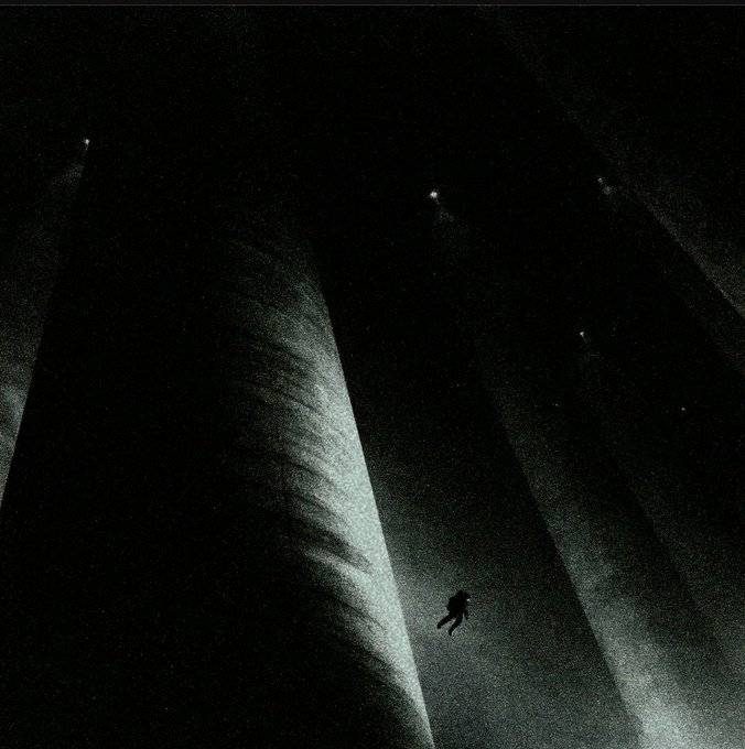 A dark, atmospheric photograph showing a tiny silhouetted figure floating or falling in a vast shadowy space illuminated by sparse spotlights from above.