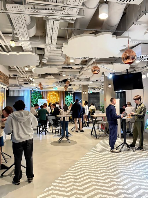 A modern indoor event space features people mingling around high tables, with cloud-shaped ceiling decorations and copper pendant lights.
