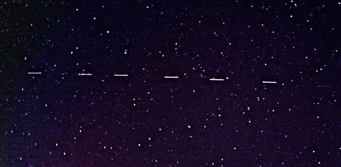 A photograph of a dark, starry night sky showing a line of evenly spaced dots moving across the frame, likely indicating satellites or a Starlink train.