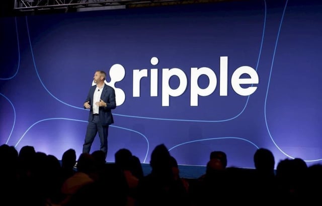 A man in a dark suit stands on stage presenting in front of a large blue backdrop displaying the 'ripple' logo, with audience members visible in the foreground.
