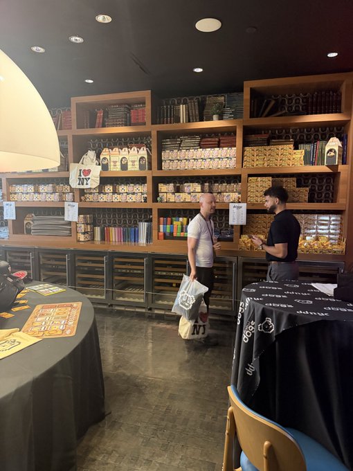 Interior of a retail shop with wooden shelves stocked with packaged goods; two men stand near the counter, one holding an 'I ❤ NY' tote bag and shopping bag.
