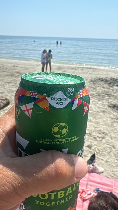 A hand holding a green Carlsberg beer can on a sandy beach, with text reading 'Deschide AICI', 'BY APPOINTMENT TO THE FOOTBALL FANS OF THE WORLD', and 'FOOTBALL TOGETHER', with the ocean and two people visible in the background.