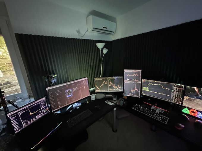 A multi-monitor trading setup with four screens displaying financial charts sits on a black desk in a dimly lit room with dark acoustic panels and an air conditioner on the wall.
