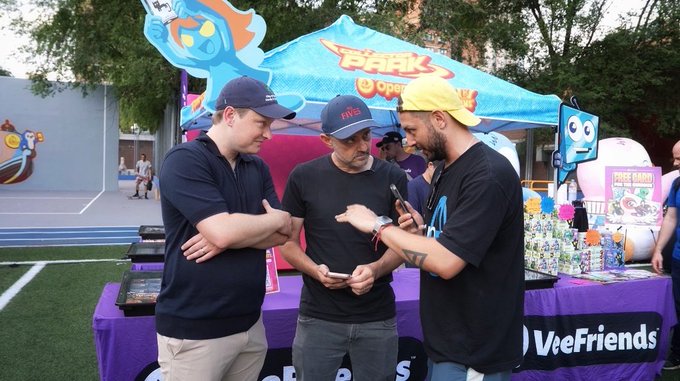 Three men standing in front of a 'VeeFriends' booth with a 'Park' sign in the background, examining a phone together.
