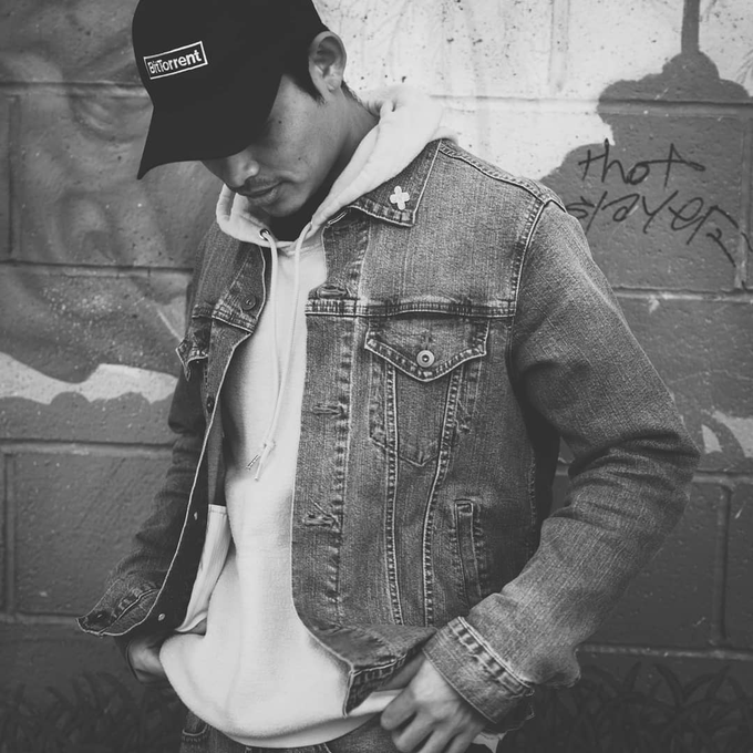 A black and white photo shows a young man wearing a denim jacket over a hoodie and a black cap with the text 'BitTorrent', standing against a brick wall with graffiti reading 'thot prayer'.